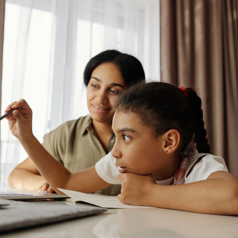 A tutor sitting beside a young girl, helping her with schoolwork at home